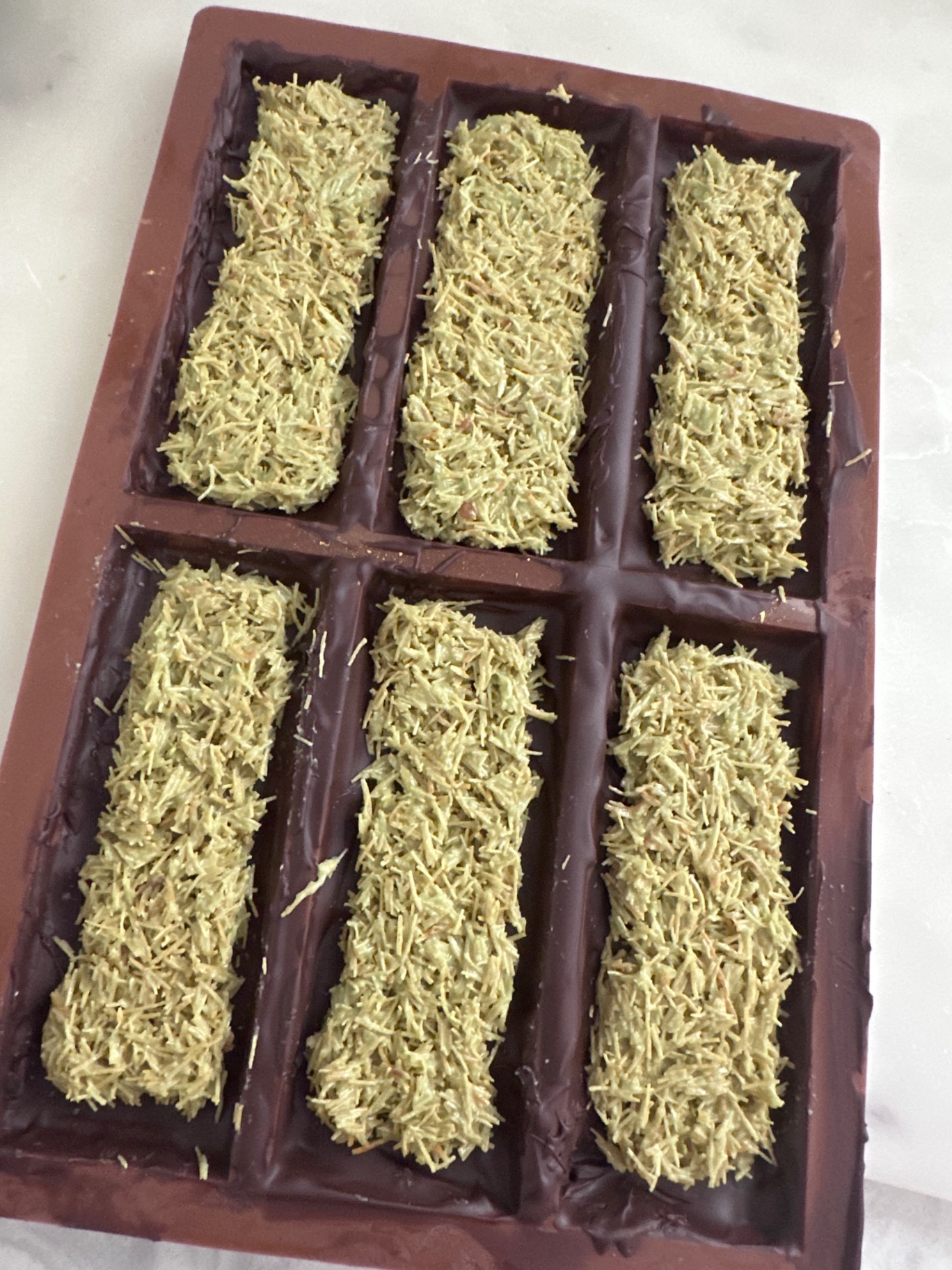 Trays of pistachio kataifi filling being layered into chocolate molds